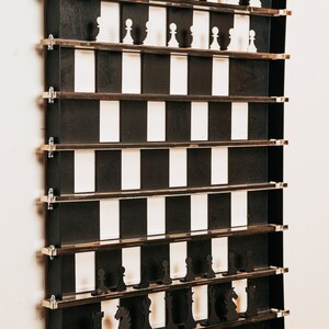May include: A black metal wall-mounted chessboard with white and black squares. The chess pieces are made of wood and are displayed on shelves. The board is labeled "Chess" and has a small tag with the logo "Architects of Fun".