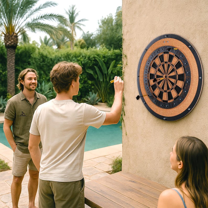 Op de afbeelding: Een dartbord met een zwart en bruin ontwerp is aan een muur bevestigd. Het dartbord heeft een kurken rand en een zwart frame. Een persoon gooit een gele dartpijl naar het bord. Twee andere mensen kijken naar het spel.
