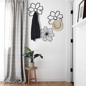 May include: Black metal flower-shaped wall hooks with a hat and jacket hanging on them. The hooks are mounted on a white wall with a white door and a gray curtain in the background.