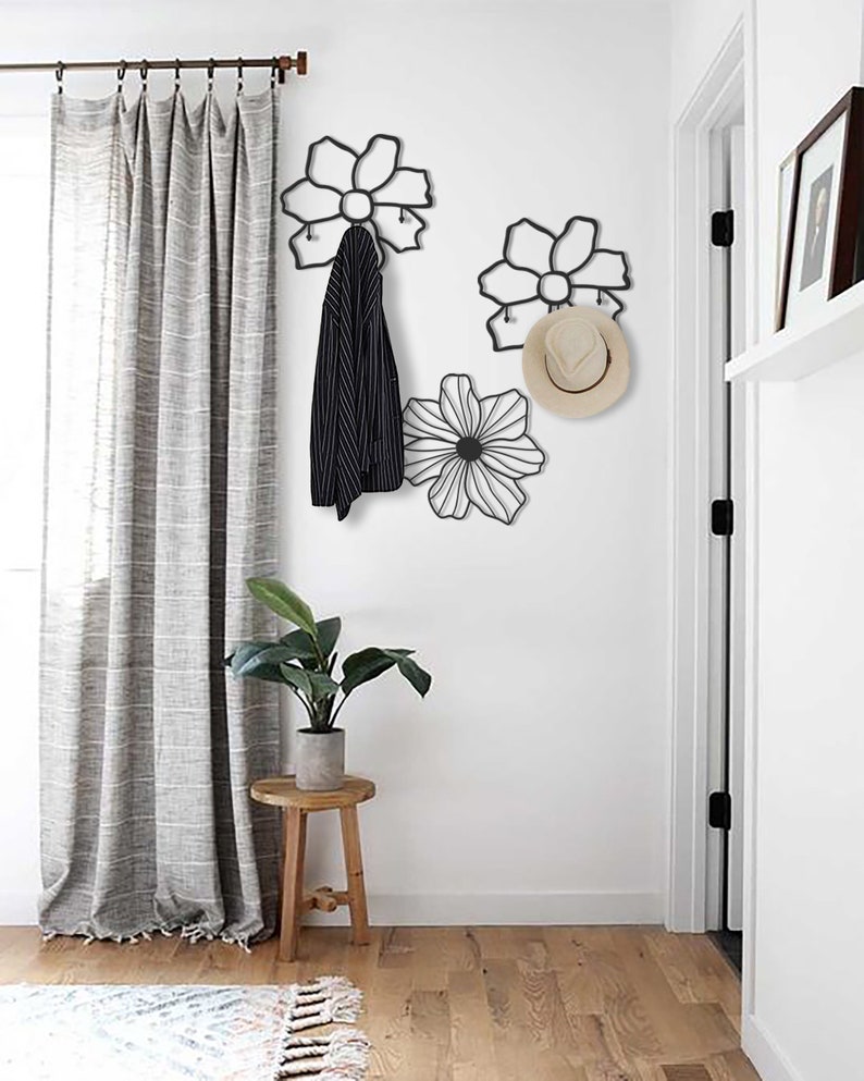 May include: Black metal flower-shaped wall hooks with a hat and jacket hanging on them. The hooks are mounted on a white wall with a white door and a gray curtain in the background.