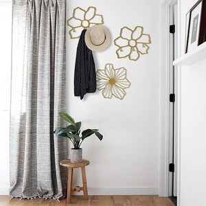 May include: Three gold metal flower-shaped wall hooks with a hat and jacket hanging on one of the hooks. The hooks are mounted on a white wall in a hallway with a wooden floor and a gray curtain.