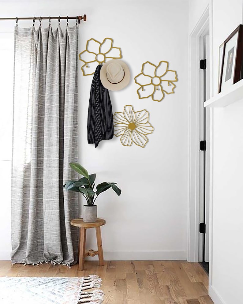 May include: Three gold metal flower-shaped wall hooks with a hat and jacket hanging on one of the hooks. The hooks are mounted on a white wall in a hallway with a wooden floor and a gray curtain.