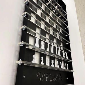 May include: A black wall-mounted chessboard with clear acrylic shelves holding white chess pieces. The board has a black and white checkered pattern and the word "CHESS!" is cut out of the black background.
