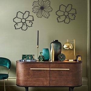 May include: Three black metal wall decor pieces in the shape of flowers. The flowers are hanging on a sage green wall above a wooden dresser with gold accents. The dresser has a green vase, a gold vase, a green glass bottle, and a gold clock on top.