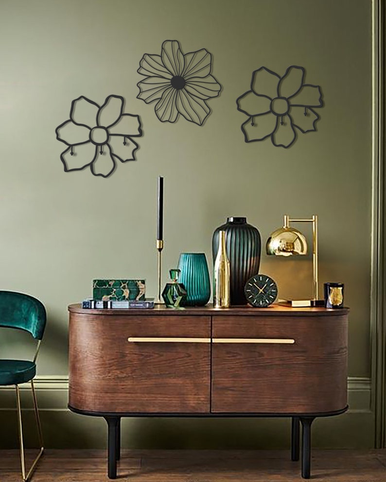 May include: Three black metal wall decor pieces in the shape of flowers. The flowers are hanging on a sage green wall above a wooden dresser with gold accents. The dresser has a green vase, a gold vase, a green glass bottle, and a gold clock on top.