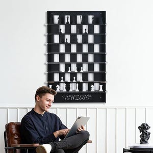 May include: A black and white chessboard wall shelf with the word "Chess" printed on the bottom. The shelf has 8 rows and 8 columns, with each square able to hold a chess piece. A person is sitting in a chair in front of the shelf, looking at a tablet.