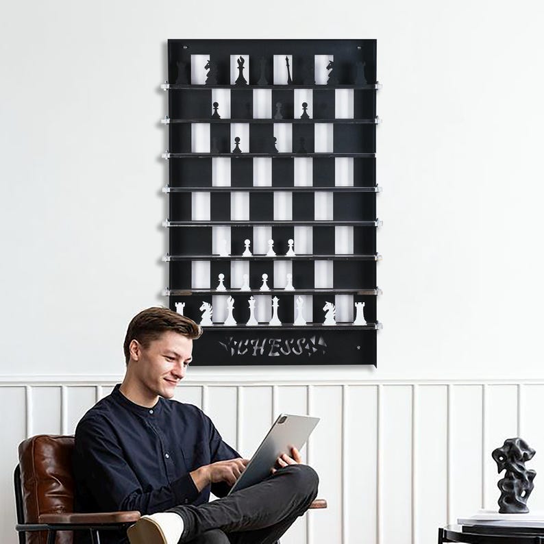 May include: A black and white chessboard wall shelf with the word "Chess" printed on the bottom. The shelf has 8 rows and 8 columns, with each square able to hold a chess piece. A person is sitting in a chair in front of the shelf, looking at a tablet.