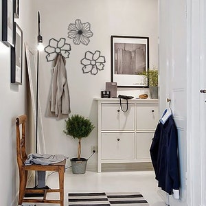 May include: Three black metal flower-shaped wall hooks with a coat hanging on one of the hooks. The hooks are mounted on a white wall in a hallway with a white dresser and a small potted plant.