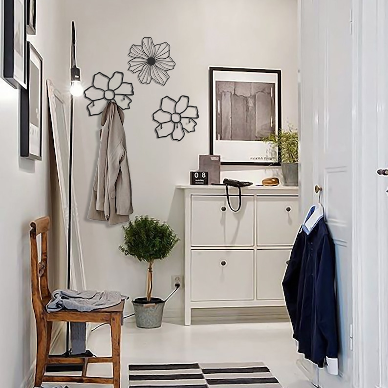 May include: Three black metal flower-shaped wall hooks with a coat hanging on one of the hooks. The hooks are mounted on a white wall in a hallway with a white dresser and a small potted plant.