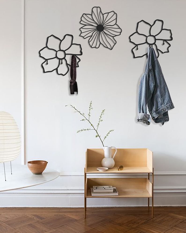 May include: Three black metal flower-shaped wall hooks with a denim jacket hanging on one of the hooks. The hooks are mounted on a white wall. A small wooden shelf with a vase and a book is on the right side of the image.