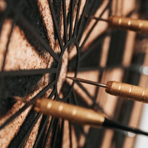 Op de afbeelding: Close-up van een dartbord met drie darts. Het bord is gemaakt van kurk en heeft een zwart metalen frame. De darts hebben goudkleurige barrels en zwarte flights, met &eacute;&eacute;n dart die de roos raakt.