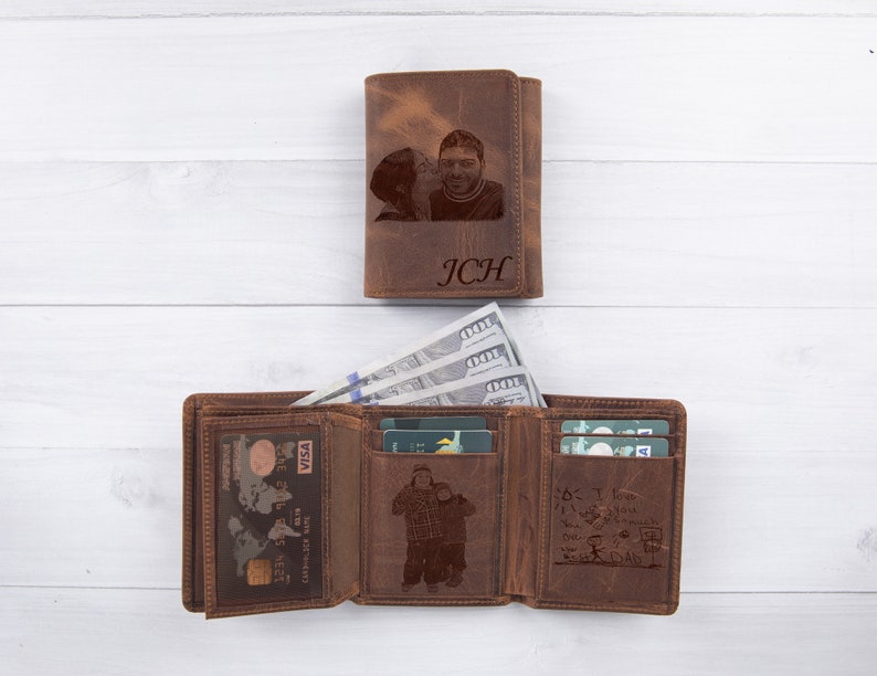 Puede incluir: Cartera de cuero marr&oacute;n de tres pliegues con una foto de una pareja grabada en la parte delantera. La cartera tiene m&uacute;ltiples ranuras para tarjetas y un clip para billetes. El texto "JCH" tambi&eacute;n est&aacute; grabado en la parte delantera.