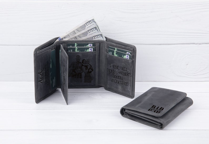 May include: Two gray leather wallets with a personalized inscription. One wallet has a photo of a family engraved on the front. The other wallet has the word "Dad" engraved on the front.