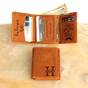 May include: A brown leather trifold wallet with a personalized inscription that reads "For Forever Kate". The wallet has a separate card slot with the inscription "Dad - You are the best" and a mustache graphic. The wallet is closed and sitting on a light brown surface.