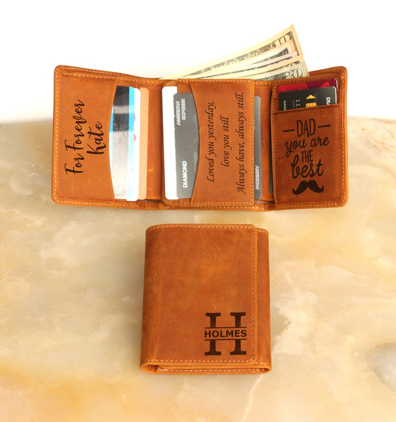 May include: A brown leather trifold wallet with a personalized inscription that reads "For Forever Kate". The wallet has a separate card slot with the inscription "Dad - You are the best" and a mustache graphic. The wallet is closed and sitting on a light brown surface.