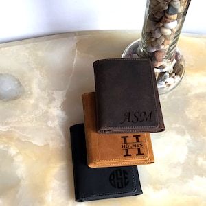 May include: Three leather wallets stacked on top of each other. The top wallet is brown with the initials "ASM" engraved on it. The middle wallet is tan with the initials "H" and the word "HOLMES" engraved on it. The bottom wallet is black with a circle monogram engraved on it.