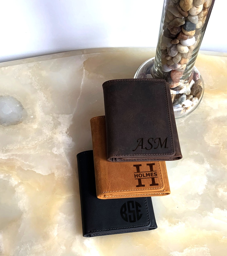 May include: Three leather wallets stacked on top of each other. The top wallet is brown with the initials "ASM" engraved on it. The middle wallet is tan with the initials "H" and the word "HOLMES" engraved on it. The bottom wallet is black with a circle monogram engraved on it.