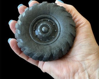 May include: A dark gray, miniature tire with a detailed tread pattern and a central hub with multiple bolts. The tire is held in a hand against a black background. The tire's diameter is approximately 3 inches.
