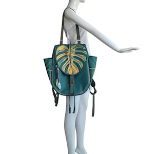 May include: A teal shoulder bag featuring a large, textured monstera leaf design in shades of yellow and green on the front flap. The bag includes two side pockets, thin shoulder straps, and decorative hanging leaf charms with a drawstring closure.