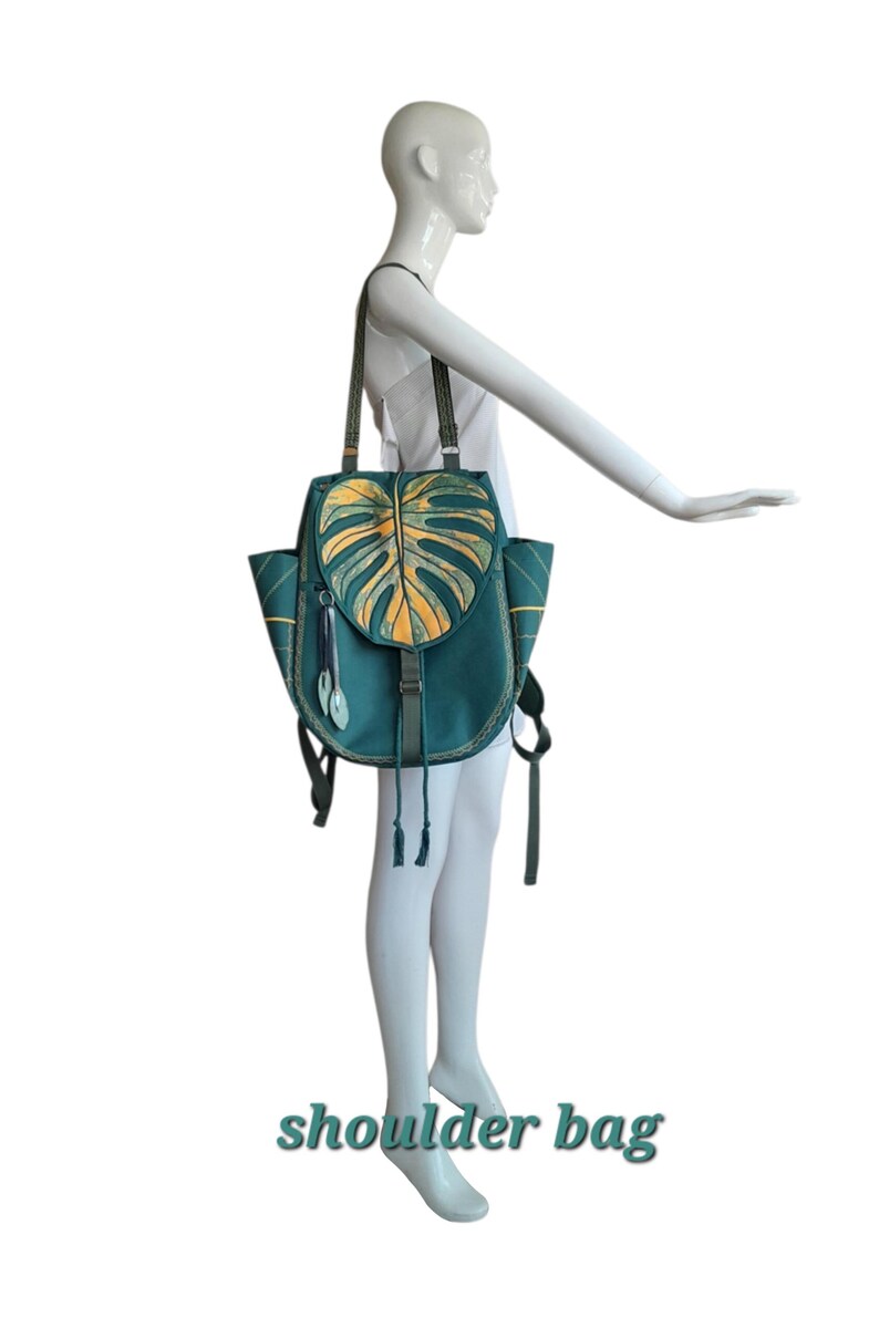 May include: A teal shoulder bag featuring a large, textured monstera leaf design in shades of yellow and green on the front flap. The bag includes two side pockets, thin shoulder straps, and decorative hanging leaf charms with a drawstring closure.