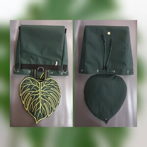 May include: A dark green backpack featuring a large, heart-shaped leaf flap with yellow embroidered veins. The bag includes a black horizontal strap, metal D-rings, a diagonal zippered pocket, and a drawstring closure with a magnetic snap button.