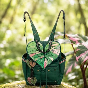 May include: A dark green backpack featuring a large heart-shaped flap designed to resemble a variegated leaf with pink and light green veins. The bag includes two shoulder straps, side pockets, and small leaf-shaped charms hanging from the front.