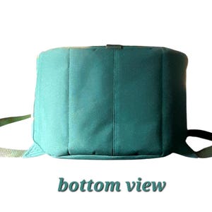 May include: A teal-colored cylindrical bag featuring a flat bottom panel and two attached fabric straps extending from the sides. The bag has a structured, vertical-seamed exterior and includes the text bottom view printed below the object.