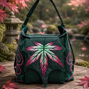 Könnte beinhalten: Eine dunkelgrüne Umhängetasche mit einer großen, zentralen Ahornblatt-Applikation mit rosa und weißem Farbverlauf. Die Tasche verfügt über einen einzelnen Tragegriff und eine dekorative Stickerei aus rosa geschwungenen Linien.