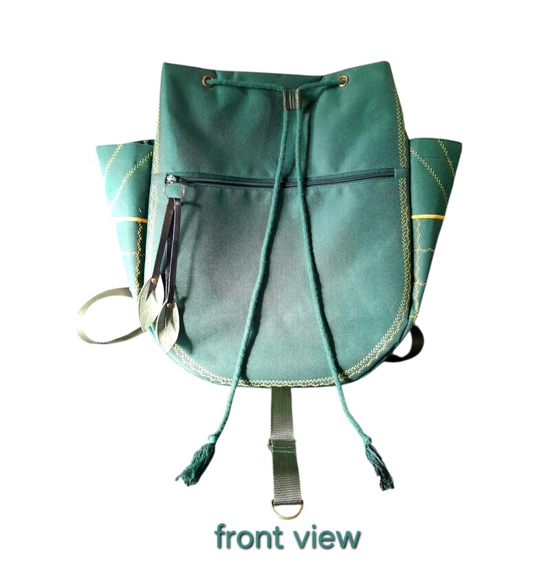 May include: A dark green drawstring backpack featuring a horizontal zippered front pocket, two side pockets with yellow stitched grid patterns, and two dangling leaf-shaped charms attached to the zipper pull. The bag has green cord straps.