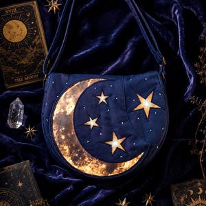 Può includere: Una borsa a tracolla blu scuro con una grande luna crescente e tre stelle a cinque punte. La luna e le stelle presentano un motivo dorato luminoso e strutturato sul tessuto blu. La borsa è dotata di una tracolla e di una patta arrotondata.