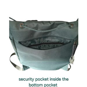 May include: A dark green backpack featuring a large front zippered pocket that opens to reveal a security pocket inside. The bag includes copper-toned metal eyelets at the top for a drawstring closure and light-colored adjustable shoulder straps.