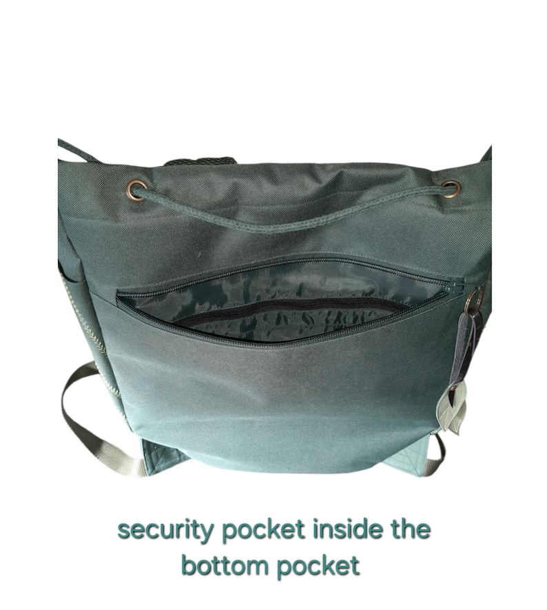 May include: A dark green backpack featuring a large front zippered pocket that opens to reveal a security pocket inside. The bag includes copper-toned metal eyelets at the top for a drawstring closure and light-colored adjustable shoulder straps.