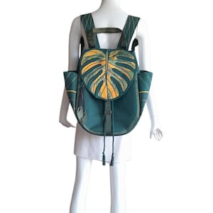 May include: A dark teal backpack featuring a prominent monstera leaf design on the flap in shades of yellow and green. The bag includes adjustable shoulder straps, side pockets, and a drawstring closure with a buckle strap, all accented with stitching.
