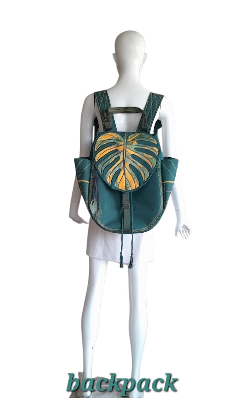 May include: A dark teal backpack featuring a prominent monstera leaf design on the flap in shades of yellow and green. The bag includes adjustable shoulder straps, side pockets, and a drawstring closure with a buckle strap, all accented with stitching.