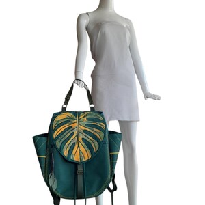 May include: A teal backpack featuring a large, yellow monstera leaf design on the front flap. The bag includes side pockets, adjustable straps, and a vertical buckle closure with long fabric ties. Two small leaf-shaped charms hang from the side.