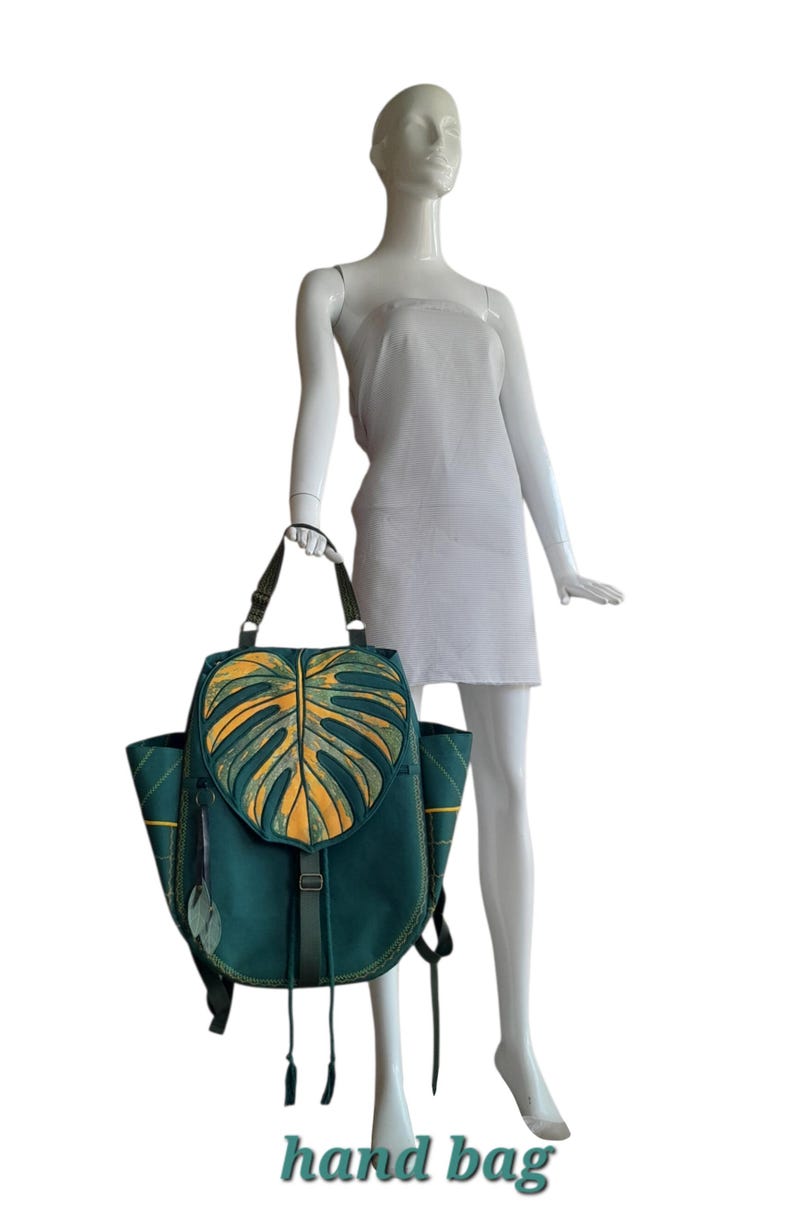 May include: A teal backpack featuring a large, yellow monstera leaf design on the front flap. The bag includes side pockets, adjustable straps, and a vertical buckle closure with long fabric ties. Two small leaf-shaped charms hang from the side.