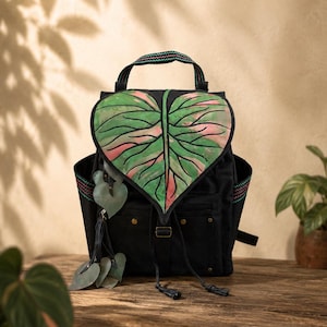 May include: A black backpack featuring a large, heart-shaped flap designed to resemble a variegated leaf with green and pink tones. The bag includes a top handle with a colorful woven pattern, side pockets, and decorative leaf-shaped charms.