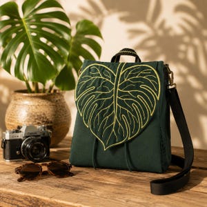 Dark green handmade convertible backpack resting on a rustic wooden table with a leaf-shaped flap embroidered in golden veins. Soft natural light, blurred monstera plant, vintage camera, and sunglasses create a cozy botanical travel aesthetic.