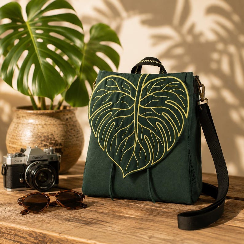 Dark green handmade convertible backpack resting on a rustic wooden table with a leaf-shaped flap embroidered in golden veins. Soft natural light, blurred monstera plant, vintage camera, and sunglasses create a cozy botanical travel aesthetic.