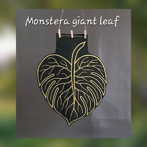 May include: A black wall hanging shaped like a Monstera leaf with gold outline detailing. The leaf is suspended by a thin twine string held by three small wooden clips. The text "Monstera giant leaf" is printed in white above the leaf.