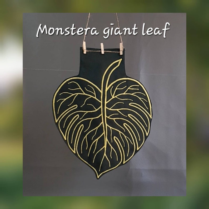 May include: A black wall hanging shaped like a Monstera leaf with gold outline detailing. The leaf is suspended by a thin twine string held by three small wooden clips. The text "Monstera giant leaf" is printed in white above the leaf.
