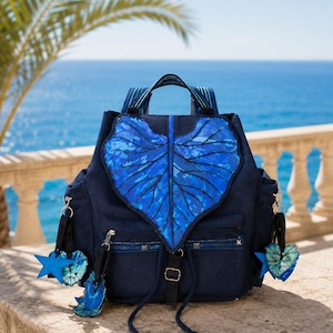 May include: A dark blue backpack featuring a large, textured leaf-shaped flap in a vibrant blue pattern. The bag includes a drawstring closure, adjustable straps, and several hanging charms shaped like stars and leaves in matching blue tones.