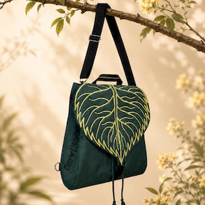 Pode incluir: Uma mochila verde-escura com uma aba grande em forma de folha com nervuras bordadas em amarelo. A bolsa tem uma alça preta ajustável, uma alça superior e um fecho de cordão com duas borlas. A palavra Convertible está impressa na lateral.