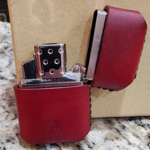 May include: A red leather lighter case with a silver metal lighter inside. The case is open, revealing the lighter's details. The case has a stitched edge and a small embossed design on the front.