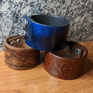 May include: Three leather wrist cuffs in varying designs and colors. One is blue with silver hardware, another is brown with a Celtic knot pattern, and the third is brown with an eagle and mountain scene.