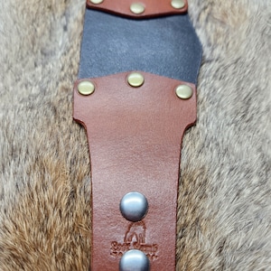 Può includere: Primo piano di un fodero per coltello in pelle con esterno in pelle marrone e interno nero. Il fodero è fissato con rivetti in ottone e bottoni automatici argentati. Il marchio "Goat Lamb Leather" è stampato sulla pelle.