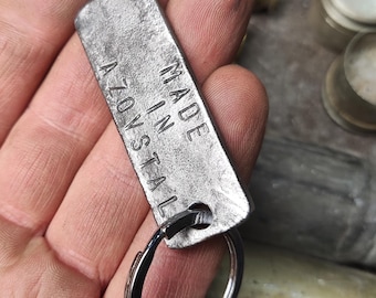 Może przedstawiać: Srebrny metalowy brelok do kluczy z prostokątnym znacznikiem z napisem "MADE IN A70V STAL".