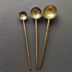Könnte beinhalten: Drei goldfarbene Löffel aus Metall mit abgerundeten Schalen. Die Löffel sind unterschiedlich groß und haben eine gehämmerte Textur.