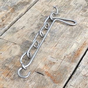 Può includere: Catena metallica color argento con ganci e anelli, appoggiata su una superficie di legno invecchiato. Il design della catena suggerisce un uso pratico. La superficie in legno ha un aspetto rustico.