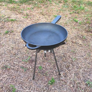 May include: A black cast iron skillet with a long handle and a small side loop handle, resting on a three-legged metal tripod stand. The skillet has a smooth, dark interior surface and a sturdy, heavy-duty construction for cooking over an open fire.